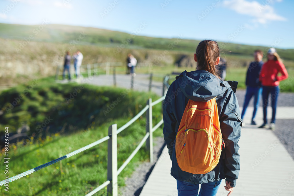 Obraz premium back view of young woman hiker hiking in beautiful mountains. female hiker with small orange packpack black lava rocks background. excursion to the national park Thingvellir in Iceland