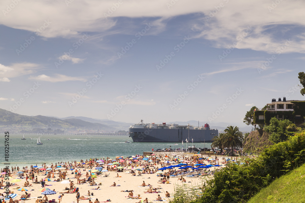 Large cargo ship leaving the harbor near a crowded beach
