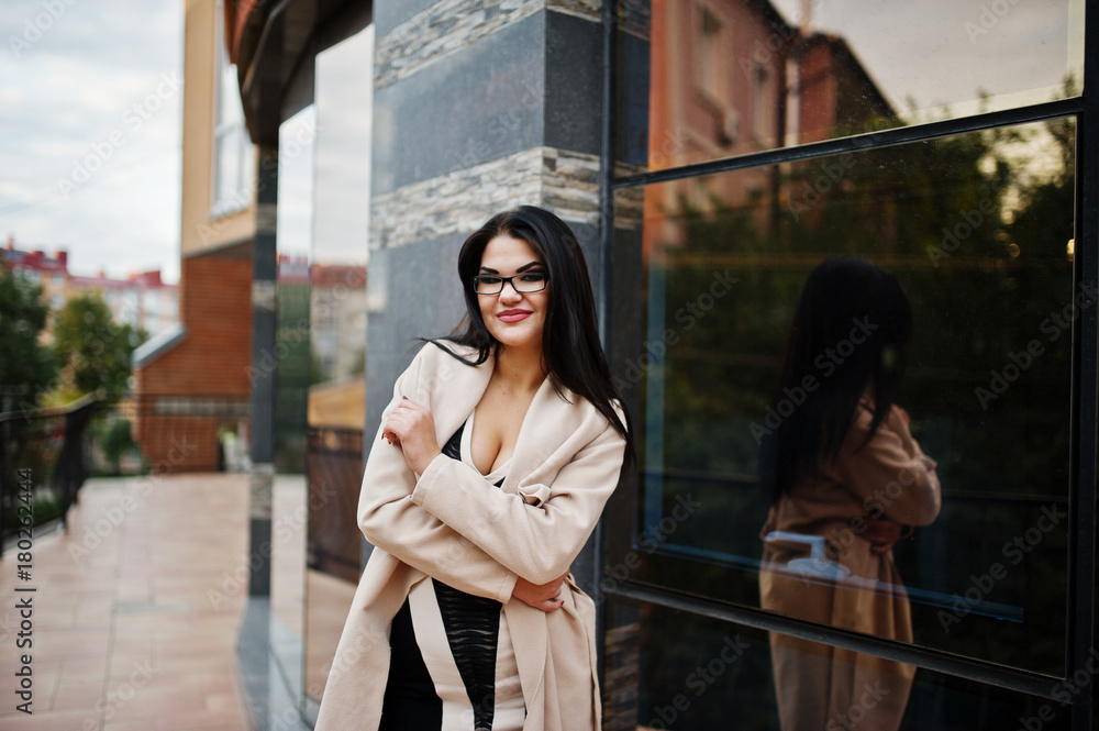 Obraz premium Black hair sexy woman in glasses and coat posed against building with modern windows.