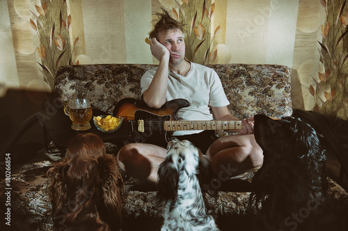 sad shaggy unkempt unshaven man with the beer and chips on the couch with a guitar , beer and chips. Three dogs setters looking at him with hungry eyes