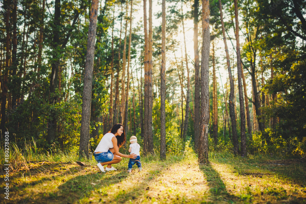 beautiful and stylish mother leads the hand of a girl of one year from birth in the park against the background of tall coniferous trees. Family values mom learns to walk child beautiful and stylish mother leads the hand of a girl of one year from birth in the park against the background of tall coniferous trees. Family values mom learns to walk child