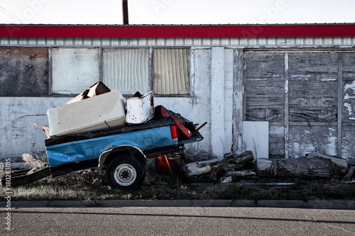 Parts of an old truck trailer and rusty appliances in front of the weathered wall of a boarded up old building, rural Kansas