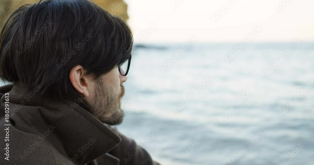 Close up of handsome young man with beard sitting on the seacoast and thinking on cloudy weather day. Outside. Portrait from back Close up of handsome young man with beard sitting on the seacoast and thinking on cloudy weather day. Outside. Portrait from back