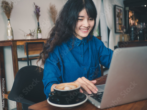 Young asian woman sitting in coffee shop at wooden table, hand holding coffee cup, Using laptop browsing internet, chatting, blogging, shopping onlie. photo