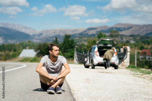 Young man is sitting in front of car on empty road. Road trip stop. Road trip break.