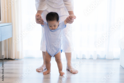 Happy Asian little baby boy learning to walk with mother help in bedroom at home. Family, child, childhood and parenthood concept