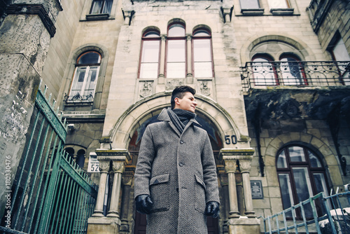 Portrait of stylish young man in warm grey coat and leather gloves on the background of old historic building.