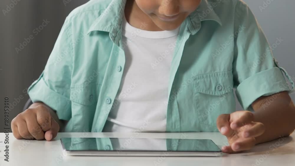 Biracial boy sitting at table with tablet, scrolling screen, modern technologies Biracial boy sitting at table with tablet, scrolling screen, modern technologies