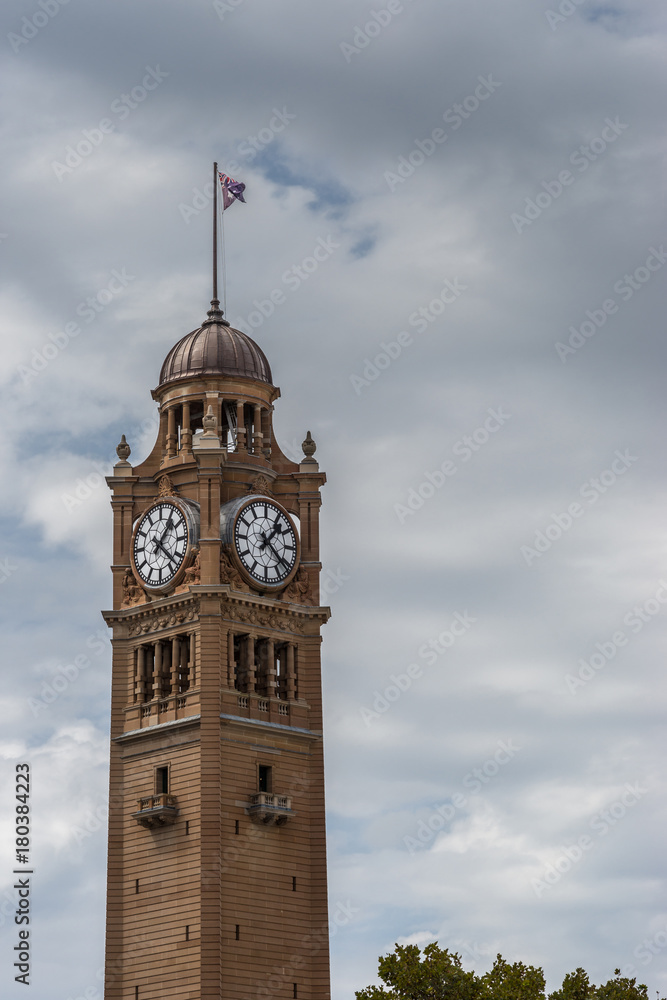 Sydney, Australia - March 25, 2017: Closeup of top of brown stone railway station clock tower against heavy sky. Flag on top.