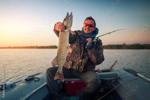 Happy amateur angler holds Pike fish sitting in the boat on the lake during sunrise
