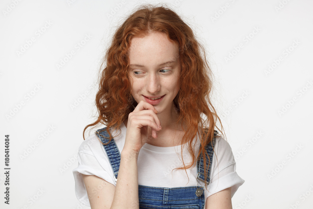Closeup headshot of young attractive European woman with red loose curly hair isolated on white background touching her chin and looking down while considering new opportunities with timid smile Closeup headshot of young attractive European woman with red loose curly hair isolated on white background touching her chin and looking down while considering new opportunities with timid smile