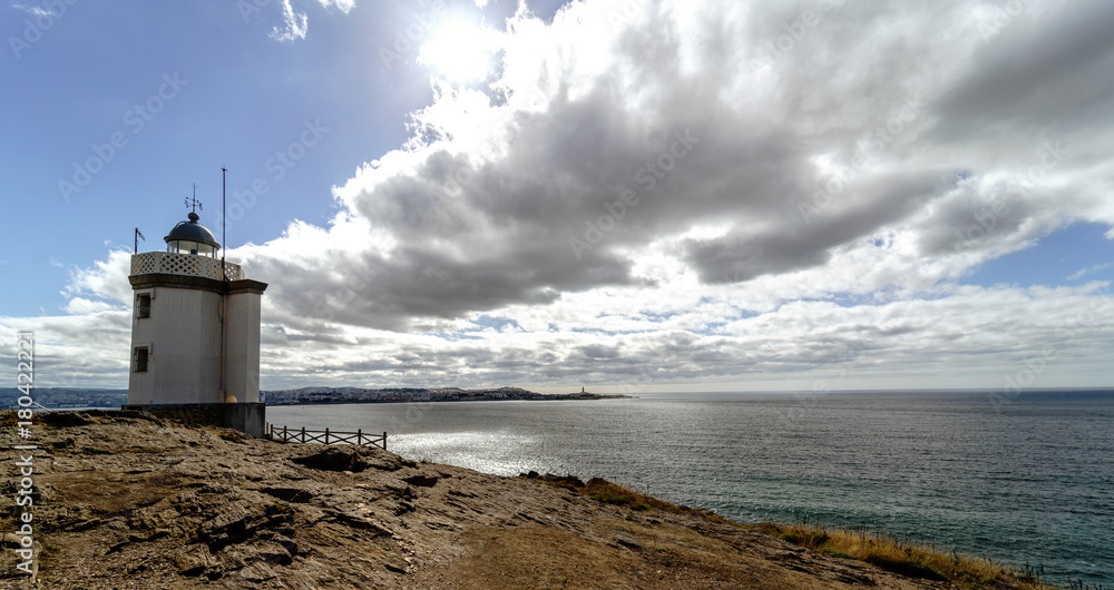 View of Mera lighthouse on a cliff on the atlantic coast of Spain in La Coruña. Sky with clouds and sun facing