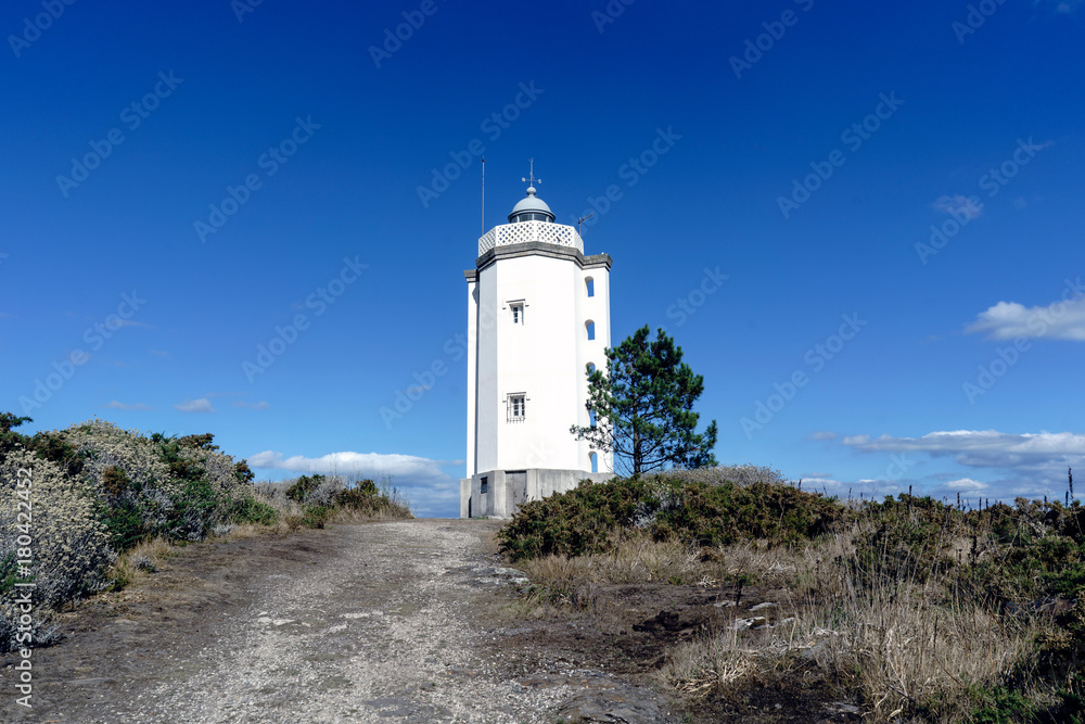 The Mera lighthouse, located on the cliffs of the coast of the Bay of La Coruna (Galicia) on the Spanish Atlantic coast. Access road. Blue sky with some clouds and calm sea The Mera lighthouse, located on the cliffs of the coast of the Bay of La Coruna (Galicia) on the Spanish Atlantic coast. Access road. Blue sky with some clouds and calm sea