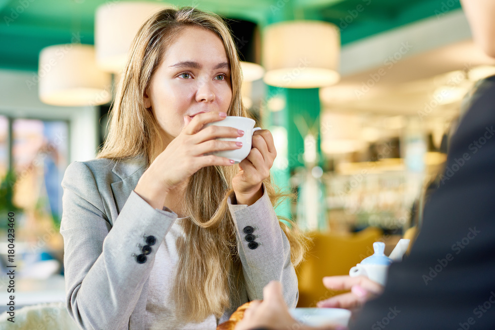 Portrait of modern young woman drinking coffee at cafe table listening to someone sitting across from her Portrait of modern young woman drinking coffee at cafe table listening to someone sitting across from her