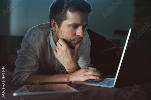 Caucasian young man lying on bed at his laptop. Working at home. photo