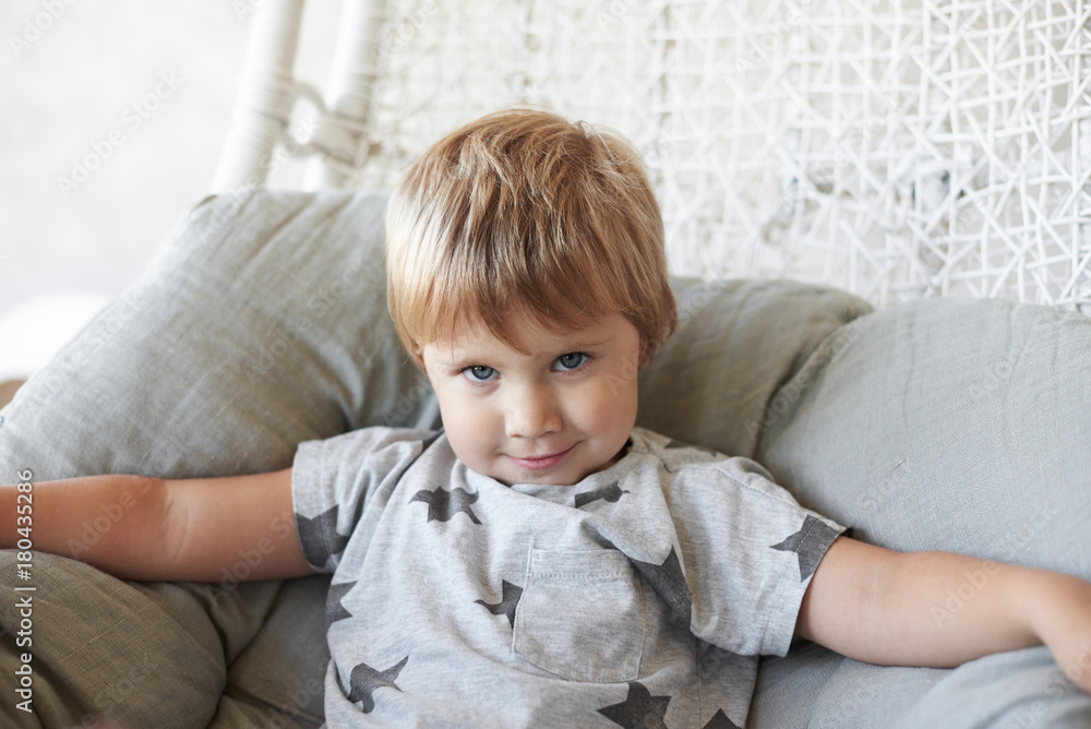 Pretty little daughter with short hairstyle and big blue eyes sitting comfortably on grey pillow in armchair, staring at camera with mysterious charming smile. Cute 5 year old girl resting at home Pretty little daughter with short hairstyle and big blue eyes sitting comfortably on grey pillow in armchair, staring at camera with mysterious charming smile. Cute 5 year old girl resting at home