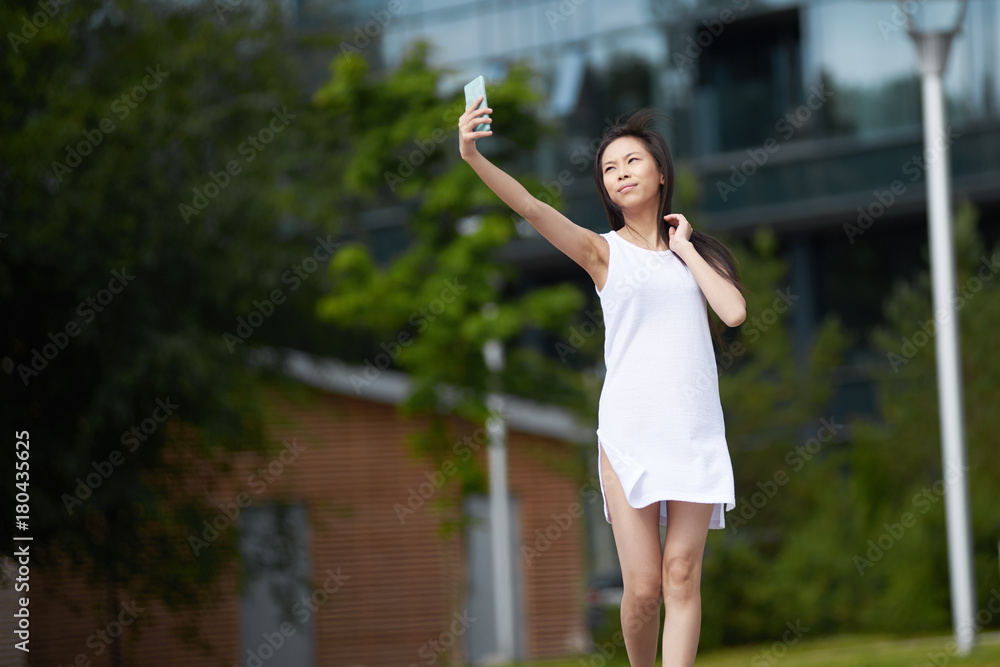 Outdoor shot of beautiful young brunette Chinese female outstretching arm with cell phone while taking selfie or having poor connection, using 3g internet to upload pictures via social networks