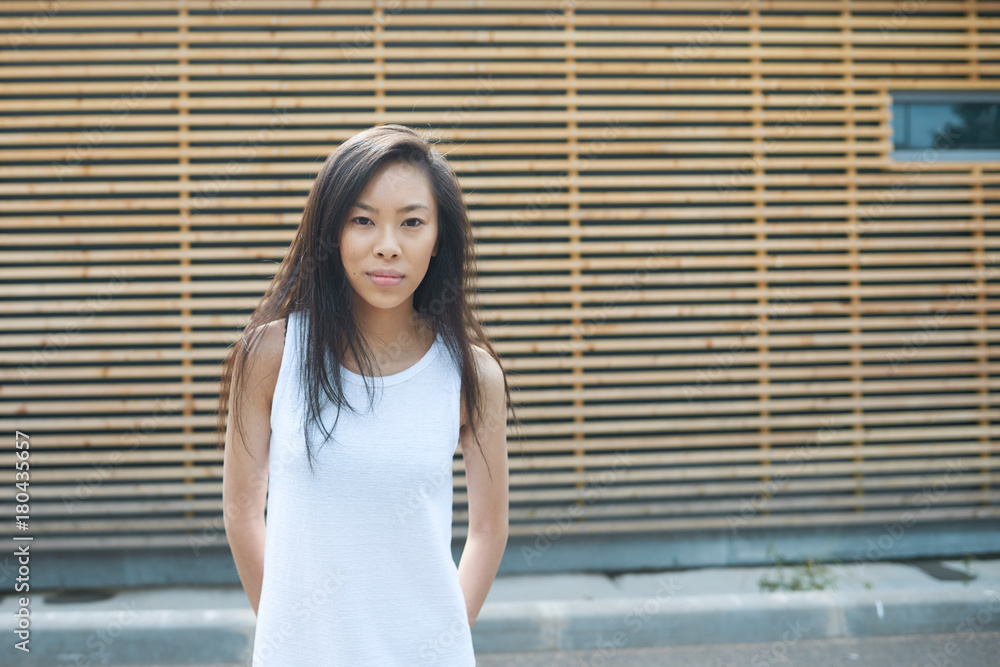 Portrait of brunette young Chinese female wearing white dress looking at camera with serious facial expression, posing at wooden building with copy space for your information or advertising content Portrait of brunette young Chinese female wearing white dress looking at camera with serious facial expression, posing at wooden building with copy space for your information or advertising content