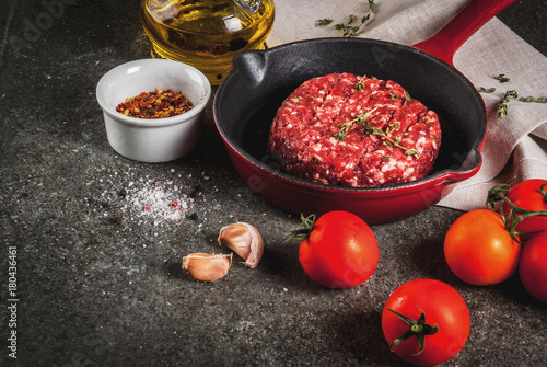 Raw organic beef meat burgers cutlets with spices, thyme, tomatoes, olive oil in frying pan on black background, copy space