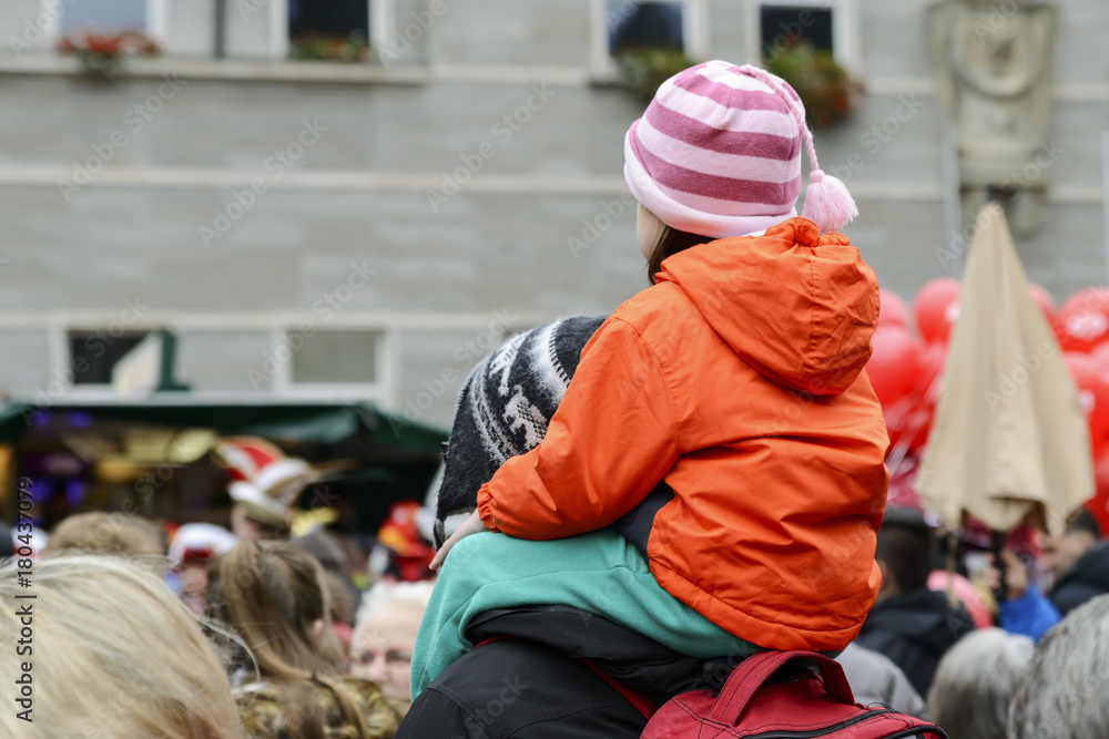 child in an orange jacket sits on his father's neck at a public event
