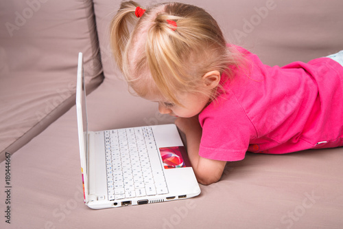 Beautiful funny blonde girl a child of two years is lying on the couch indoors and uses a white laptop computer technology with colorful prints pushing the buttons on the keyboard.