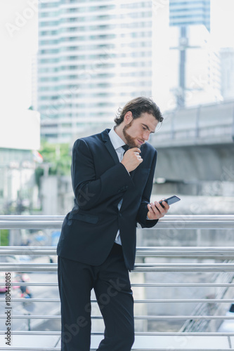 Smart businessman in full suit standing outdoors using smartphone for manage business project in cityscape background