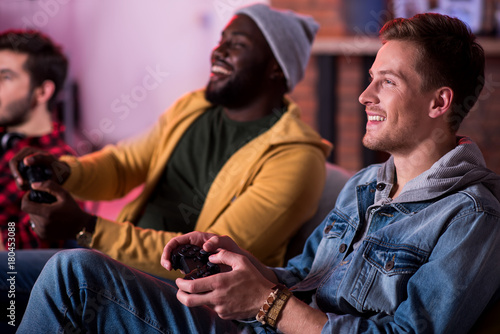 Adult party. Joyful fashionable attractive guys are enjoying play station. They are sitting on sofa and expressing gladness while holding joystick and looking at monitor. Selective focus