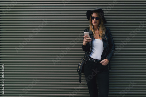 Portrait of beautiful hipster girl looking at mobile phone while chatting online while standing outdoors. Stylish student in sunglasses and hat reading messages while standing on black wall background