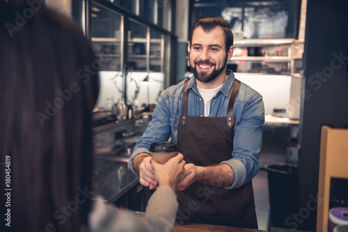 Portrait of smiling bearded worker holding out hand with mug of beverage to woman in modern cafe. Order concept