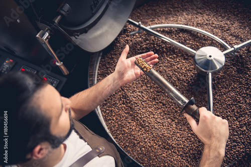 Slika na platnu Top view serious bearded worker near coffee roaster controlling level of grain roasting