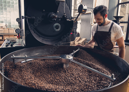 Side view smiling unshaven worker watching at coffee beans situating in grain chiller. Industry concept photo