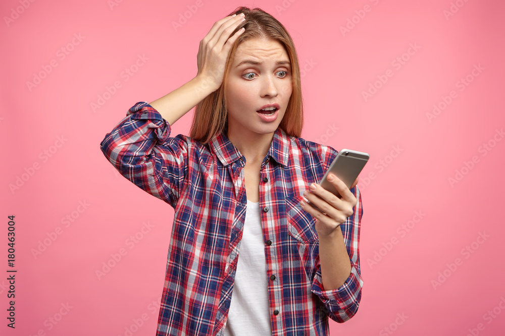 Shocked woman with long hair, keeps hands on head, looks into screen of mobile phone, recieves message remainding her about necessary thing to do, forgot about it completely, isolated on pink wall Shocked woman with long hair, keeps hands on head, looks into screen of mobile phone, recieves message remainding her about necessary thing to do, forgot about it completely, isolated on pink wall
