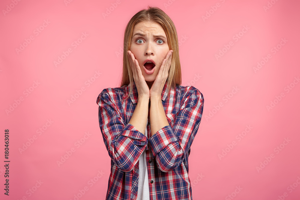 Portrait of shocked stupefied young female looks with widely opened mouth, keeps hands on cheeks, doesn`t believe in her failure, has puzzled and frustrated look, isolated over pink background Portrait of shocked stupefied young female looks with widely opened mouth, keeps hands on cheeks, doesn`t believe in her failure, has puzzled and frustrated look, isolated over pink background
