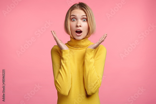 Surprised terrified female gestures with uncertainty  stares at camera  puzzled as doesn t know anwser on tricky question  isolated over pink background. People  body language  emotions concept