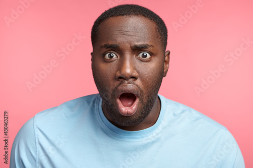 Shocked black man stares at camera, opens mouth widely, can`t believe in what he sees, being in stupor, isolated over pink background. Emotional dark skinned young plump male has terrified look