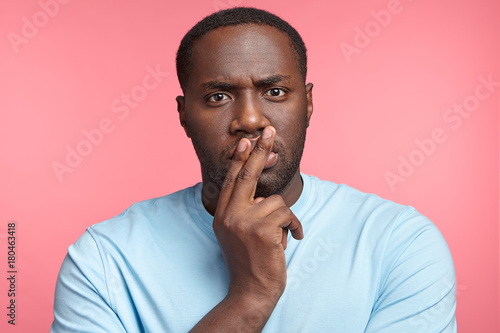 Handsome young dark skinned man looks pensively directly into camera, thinks over future plans. Male employee develops new strategy, isolatd over pink background. People and confidence concept