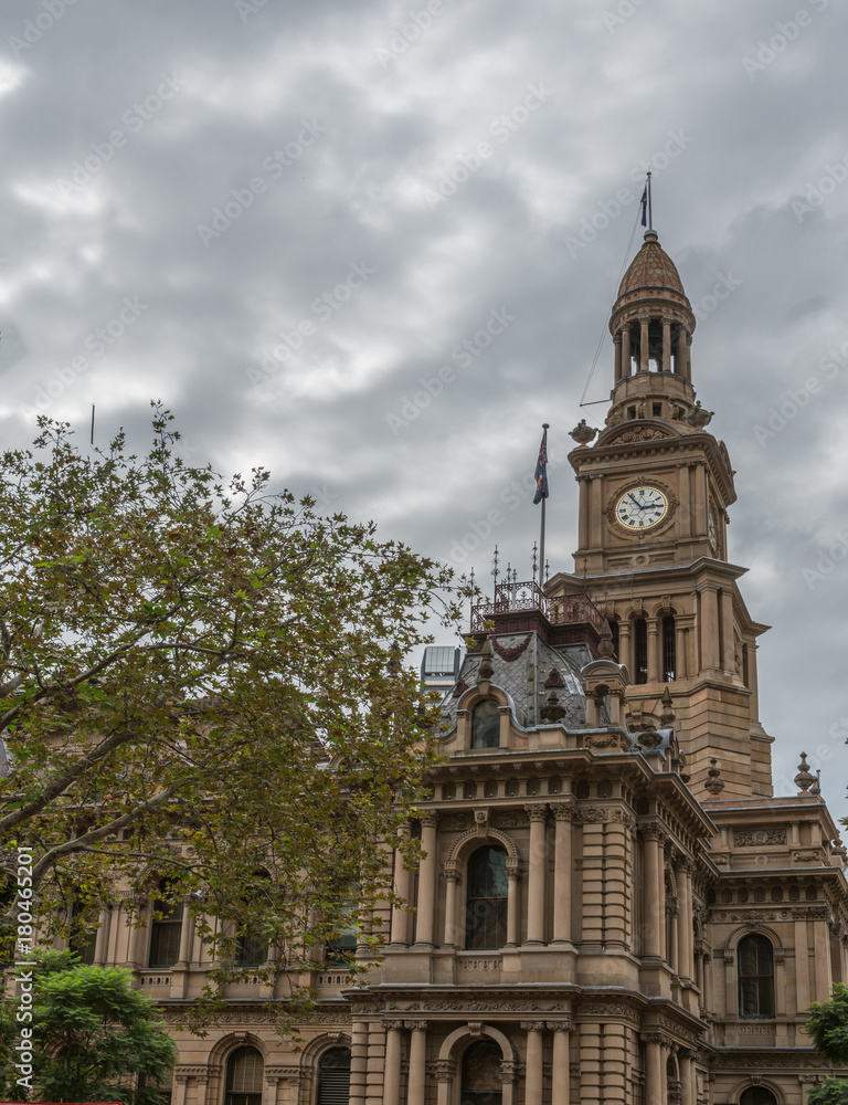 Sydney, Australia - March 25, 2017: View on corner of brown stone historic monumental City Hall with clock tower under heavy sky and tree in foreground.