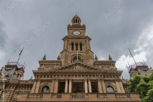 Photography Sydney, Australia - March 25, 2017: Vertical shot upwards of brown stone front facade monumental historic City Hall building under heavy sky and some green foliage on the side