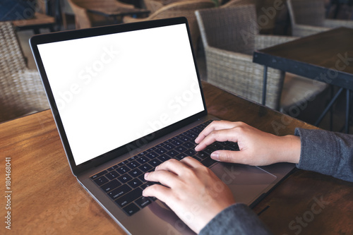 Mockup image of business woman using and typing on laptop with blank white screen and coffee cup on glass table in modern loft cafe