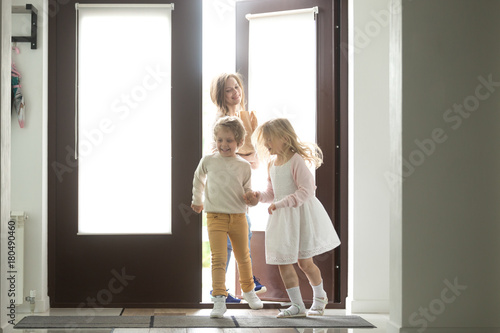 Happy young mother with son and daughter opening door coming back home from mall, single mom and small kids returning to house holding paper bag with food, family grocery shopping together concept