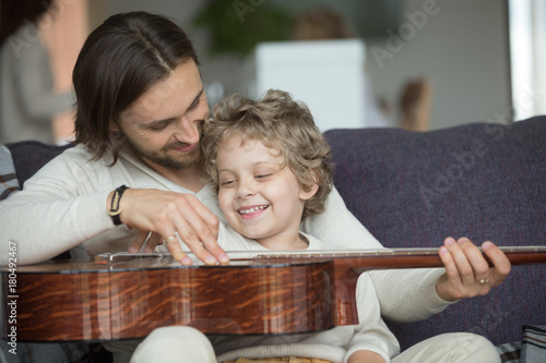 Young father teaching little son to play musical instrument at home, loving dad strumming guitar with small boy, parent and child relaxing on sofa, family leisure music activity, musically gifted kid