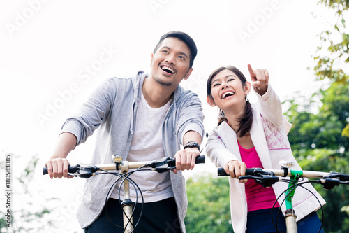 Young Asian couple laughing together while riding bicycles outdoors in summer 