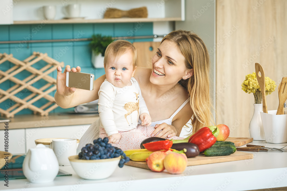 Young mother smiling, cooking and playing with her baby daughter in a modern kitchen. Healthy food concept. Working at home. Woman with a baby doing a selfie.