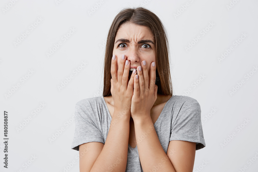 Portrait of charming unhappy beautiful brunette caucasian student girl with long hairstyle in stylish gray shirt clothing mouth with hands, looking n camera with shocked and disgust face expression Portrait of charming unhappy beautiful brunette caucasian student girl with long hairstyle in stylish gray shirt clothing mouth with hands, looking n camera with shocked and disgust face expression