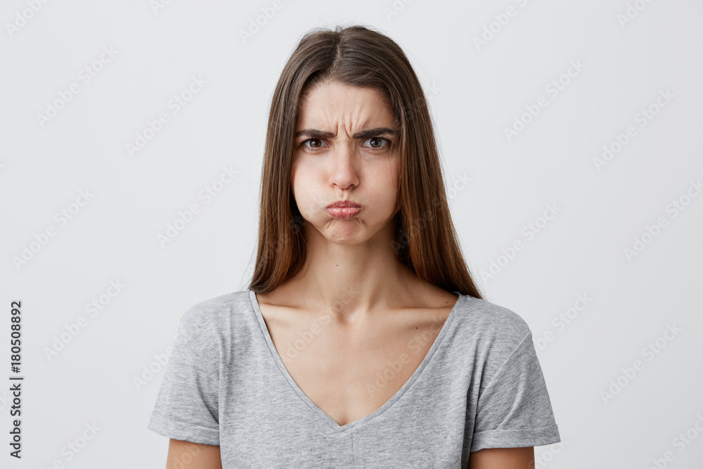 Close up of beautiful charming funny young caucasian female with dark long hair in casual gray shirt inflate cheeks, looking in camera with unsatisfied and offended face expression, having fun on Close up of beautiful charming funny young caucasian female with dark long hair in casual gray shirt inflate cheeks, looking in camera with unsatisfied and offended face expression, having fun on