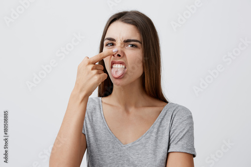 Close up portrait of funny cheerful cute caucasian girl with dark hair in fashionable outfit lifting nose with hand, showing tongue, looking in camera with silly face expression, having fun.