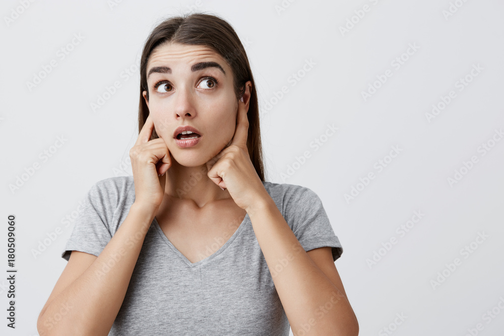 I don't hear anything. Body language. Close up of young attractive caucasian girl with long hair in casual gray t-shirt plugging ears with fingers, looking aside with raised eyebrows and surprised I don't hear anything. Body language. Close up of young attractive caucasian girl with long hair in casual gray t-shirt plugging ears with fingers, looking aside with raised eyebrows and surprised