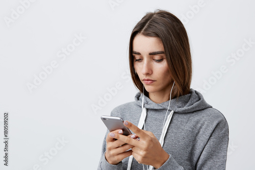Modern lifestyle. Portrait of young beautiful charming caucasian student girl with dark long hair in gray hoodie looking in smartphone with serious and calm expression, looking through social networks
