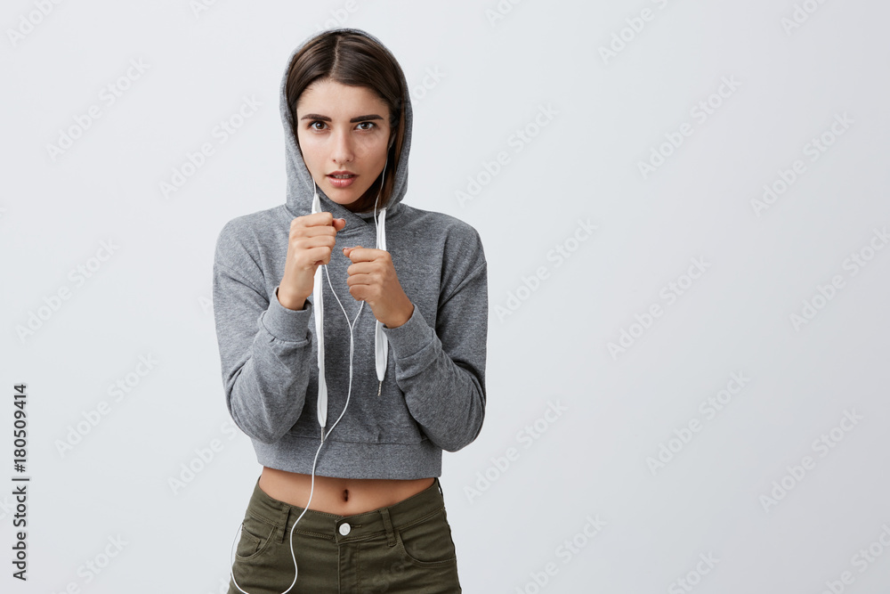 Health and beauty. Close up of young concentrated good-looking caucasian girl with dark long hair in sporty gray hoodie holding hands in fight pose, training for box competition in gym. Health and beauty. Close up of young concentrated good-looking caucasian girl with dark long hair in sporty gray hoodie holding hands in fight pose, training for box competition in gym.