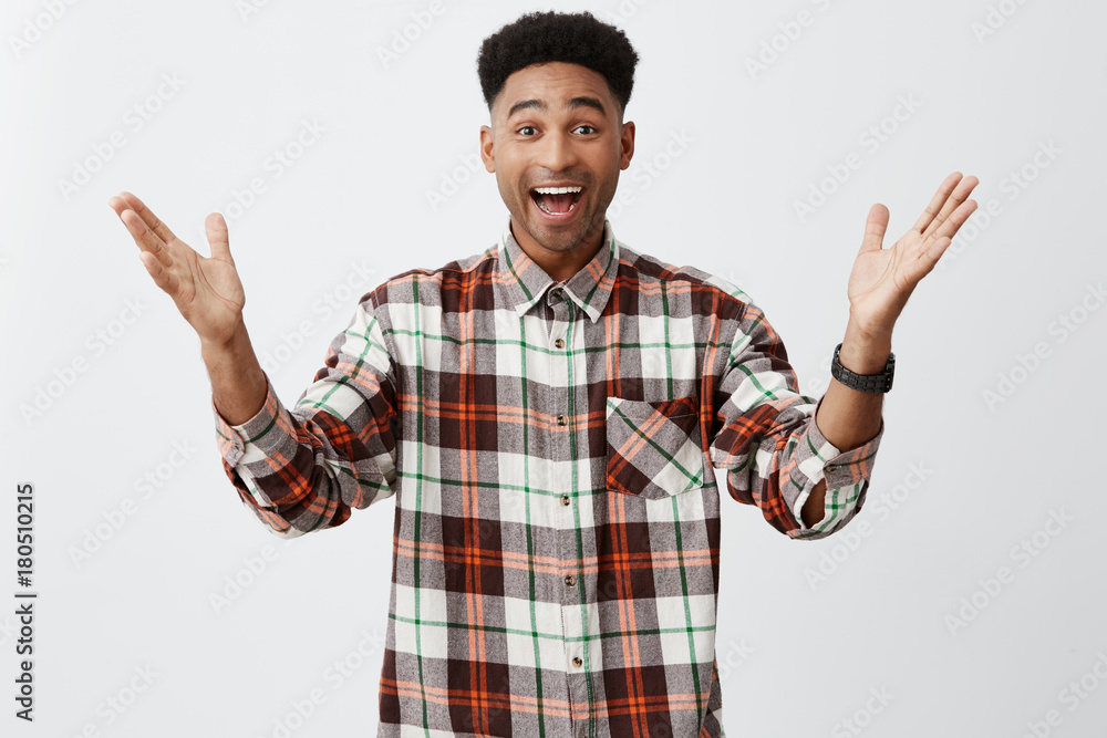 Naklejka premium Positive emotions. Isolated portrait of young happy tan-skinned guy with afro haircut in long-sleeved checkered shirt spreading hands, being excited to see old friend after vacation.